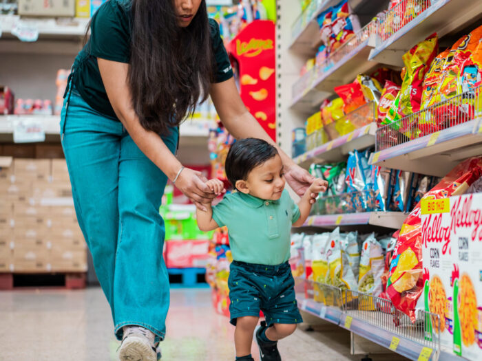 Parent supports a toddler, while they both walk along a supermarket aisle.