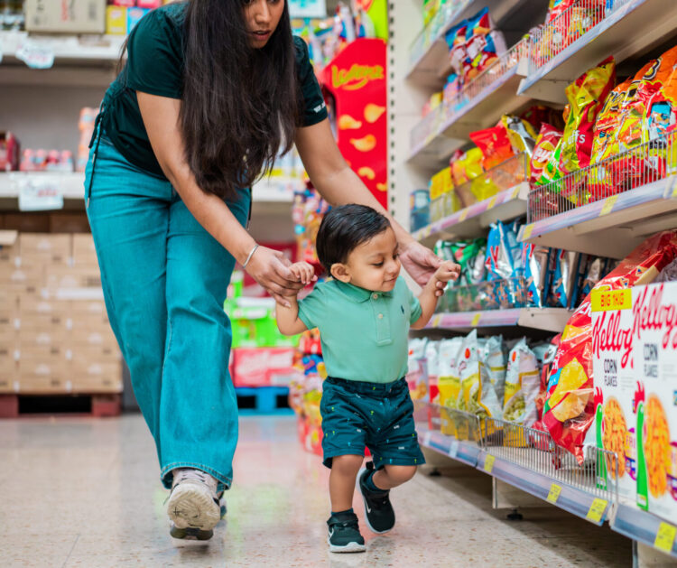 Parent supports a toddler, while they both walk along a supermarket aisle.