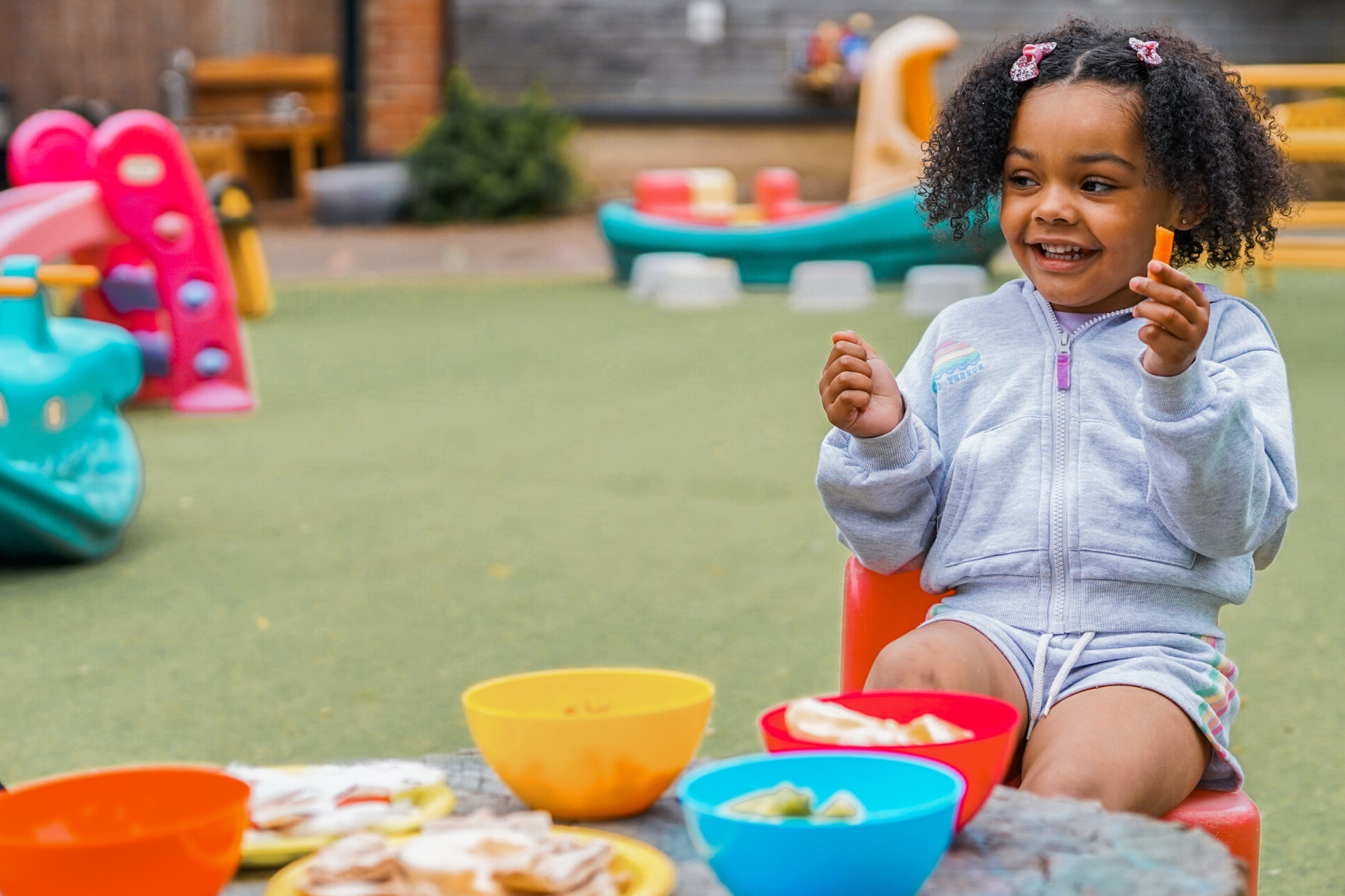 A smiling child sits in a playground, with colourful food bowls on a table in front of them.