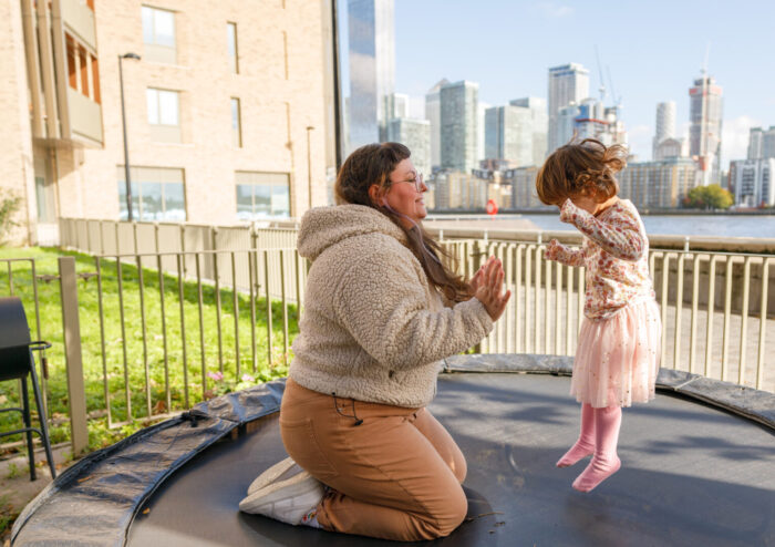 Woman kneels on trampoline with bouncing child. Photograph by Jade Reynolds-Hemmings