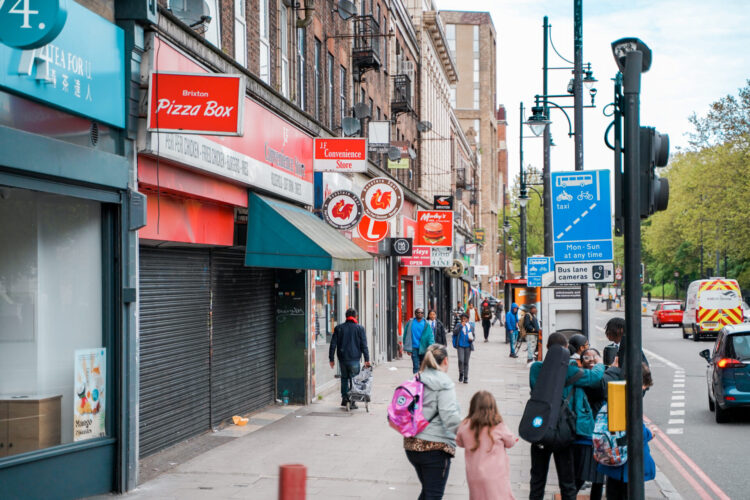 London city street with shopfronts and cars alongside walking pedestrians.