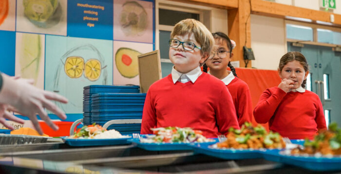 Primary school children in red jumpers wait in line for their school dinner.