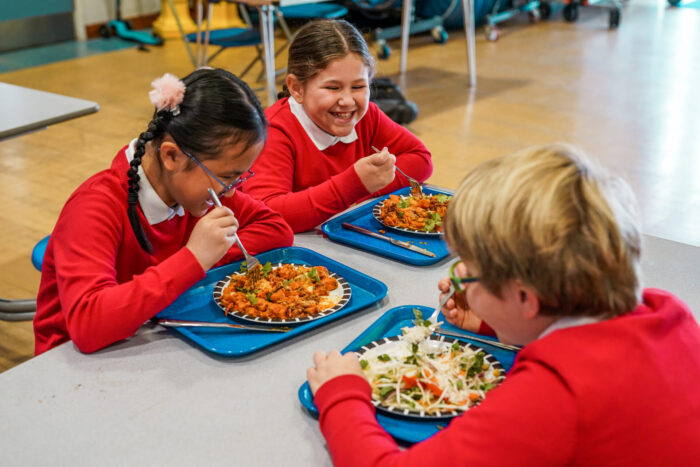 Primary school children wearing red school jumpers sit at a table, laughing together while they eat their school dinner.