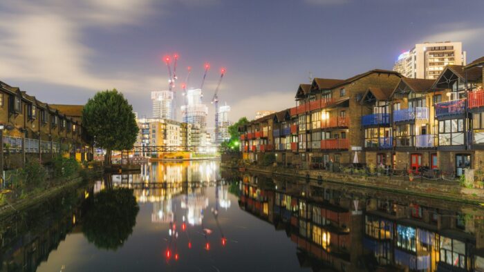 Night-time image of waterside apartment buildings, warmly lit with lights from inside, with towers and cranes lit in red in the background. Photograph by Henry Ren