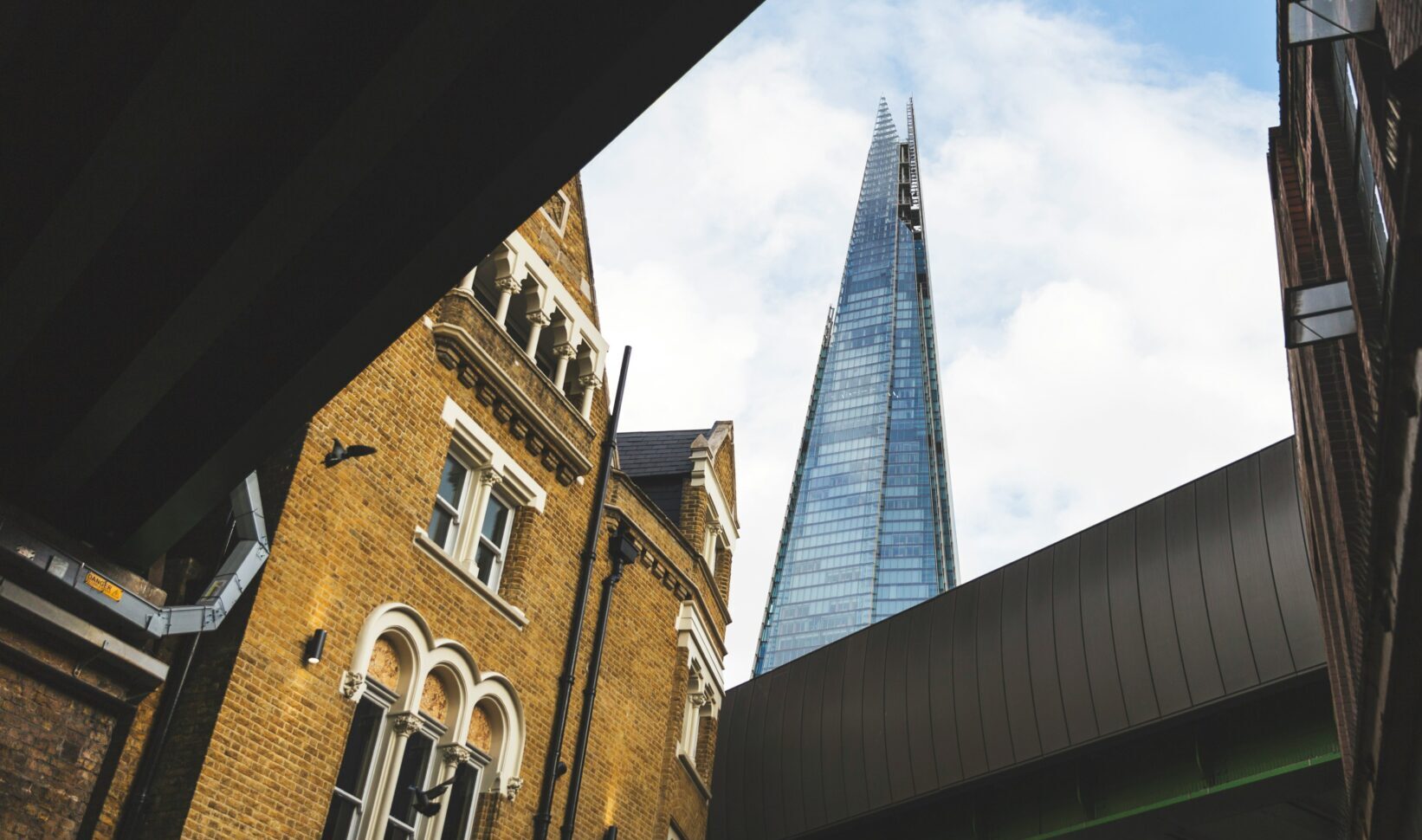 The Shard, viewed from below at street level, with yellow-brick residential buildings and bridge in foreground. Photograph by Paul Knight