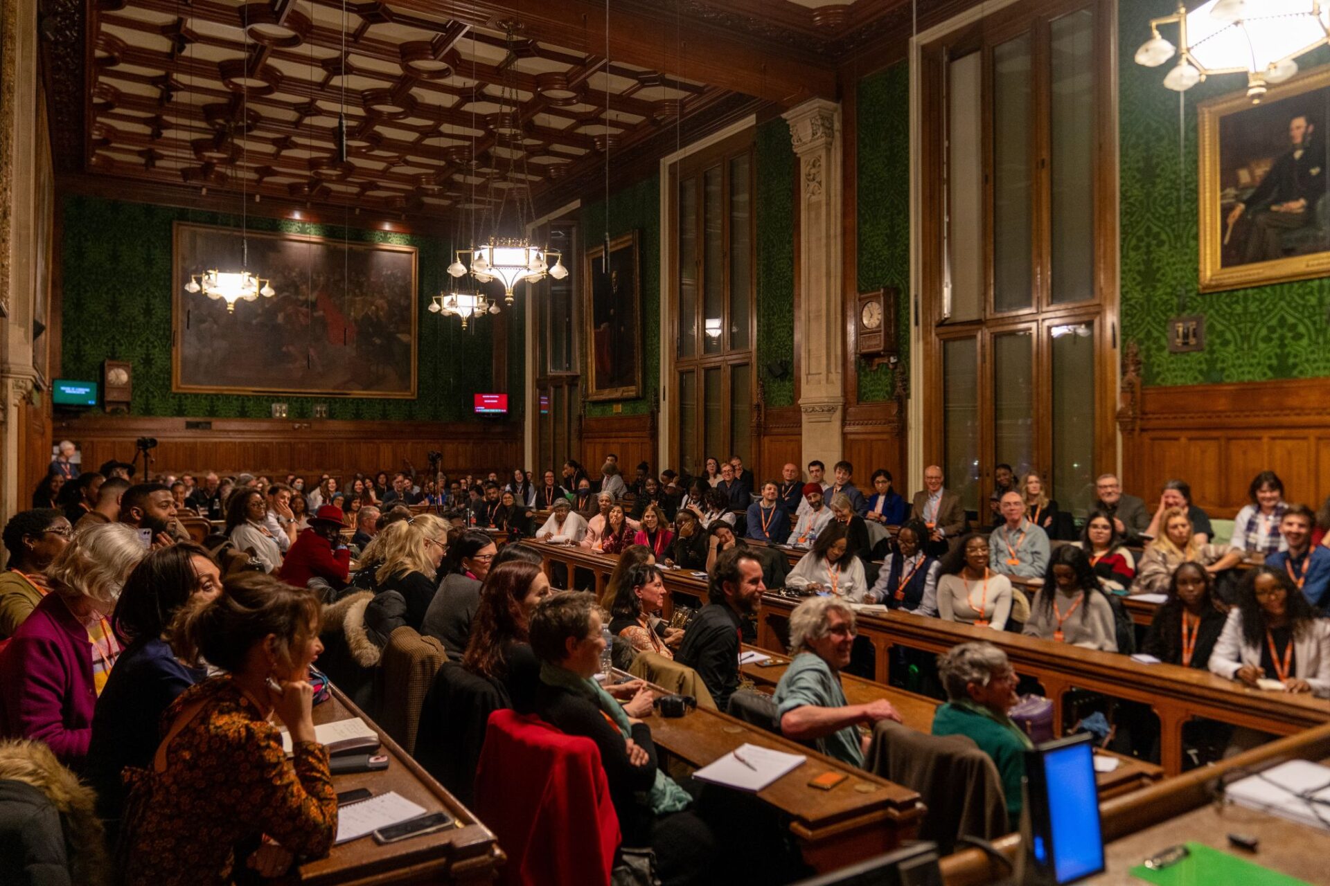 People are sitting on wooden benches and listening to a speaker at the Enact Equality event in Parliament. The room has floor to ceiling windows and portraits hung on the walls.