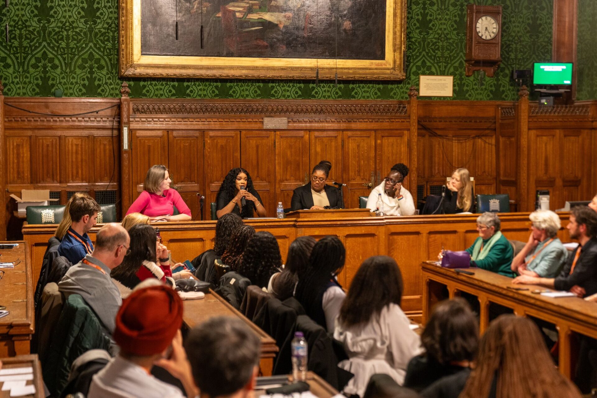 A panel of speakers at the Enact Equality event in Parliament. They are sitting on chairs at the front of the room and have microphones. In the foreground, people are sitting on wooden benches and are listening.