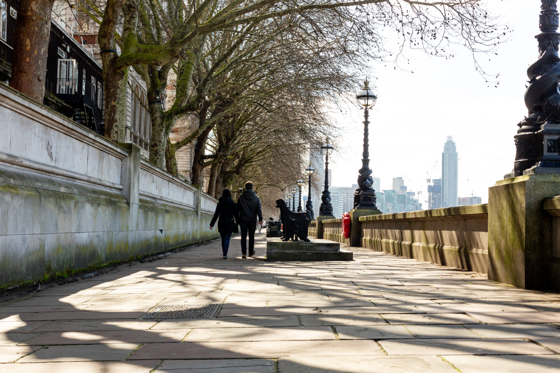 People are walking by the River Thames. The pavement is lined with trees and lamp posts. There is a bench in the distance.