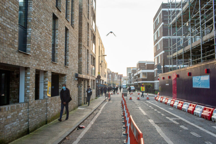 People are walking down a street. On the other side, there is scaffolding and construction works taking place, with traffic cones on the road.