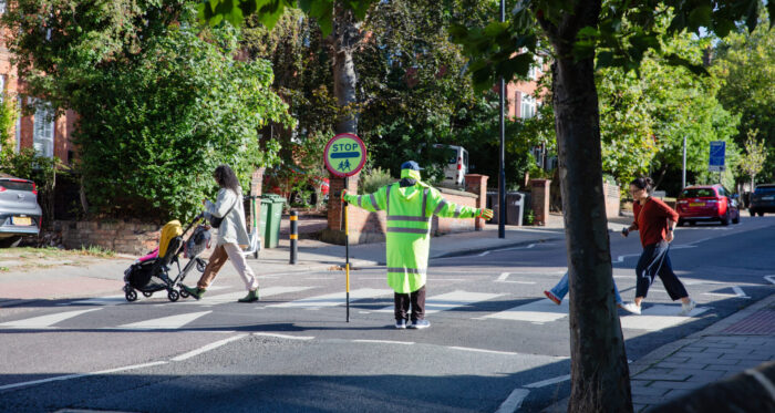 A lollipop person standing in the middle of a road. A person pushing a pushchair and another person is behind them, walking across the road.