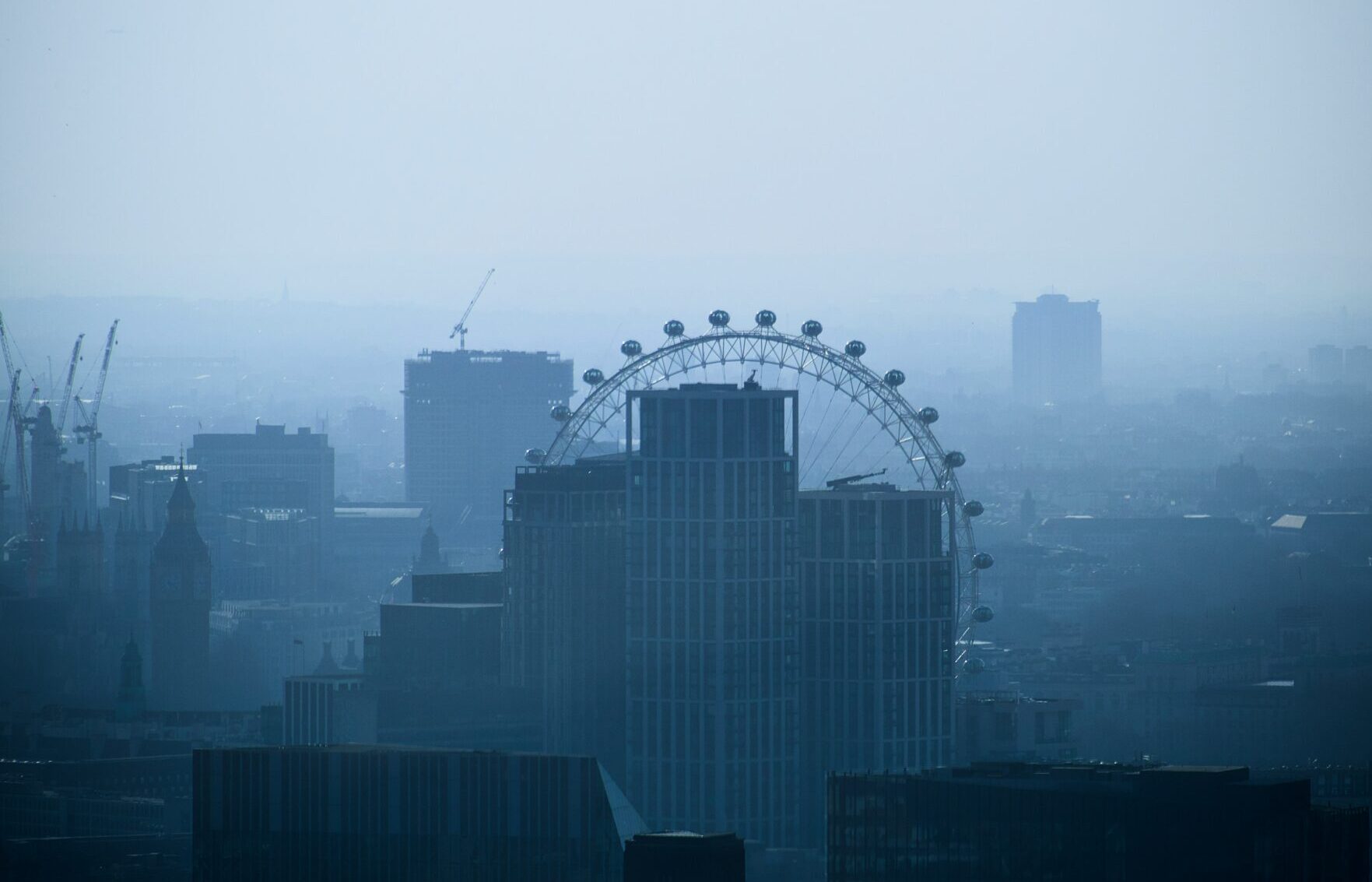 The London skyline with mist. The London eye and some tall building are visible. Photo by Ashley Gorringe via Unsplash.