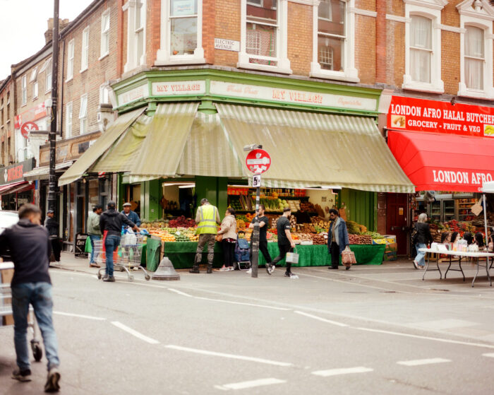 Electric Avenue in Brixton. A street corner with various shops and people walking. There is a fruit and vegetable shop with a stall outside.