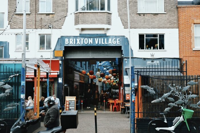 The entrance to Brixton Village. There are restaurants either side of the entrance, and people are sitting outside on tables and chairs.