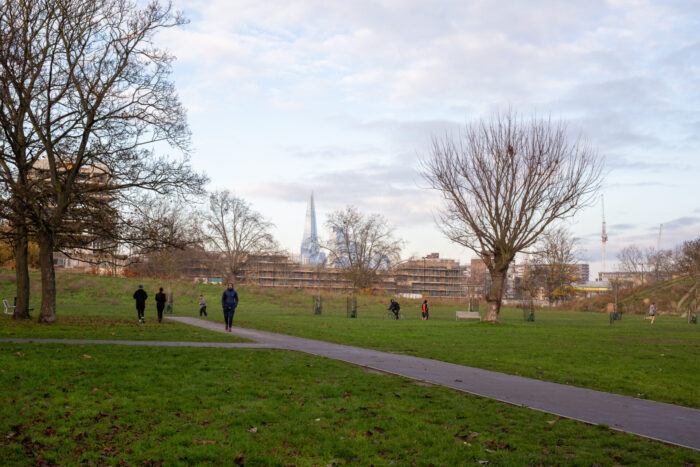 A park with trees and grass in the foreground. People are walking and running through the park. In the distance, the Shard and some buildings under construction are visible.