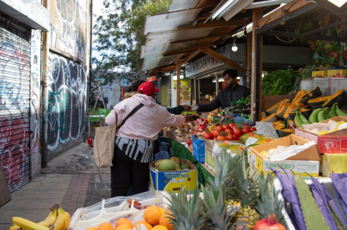 A fruit and vegetable stall with various fruits in boxes on display. People are buying produce from the stall owner.