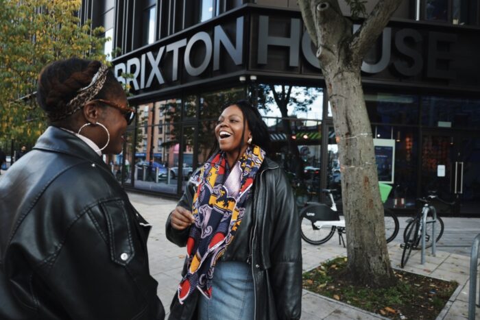 Two people are talking on a street in Brixton, London. They are standing outside of a building with a sign: 