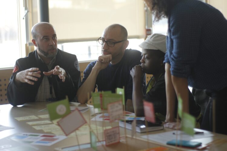 A group of people around a table are sitting and talking together, as part of a workshop. Three people are sitting around a table and one person is standing.