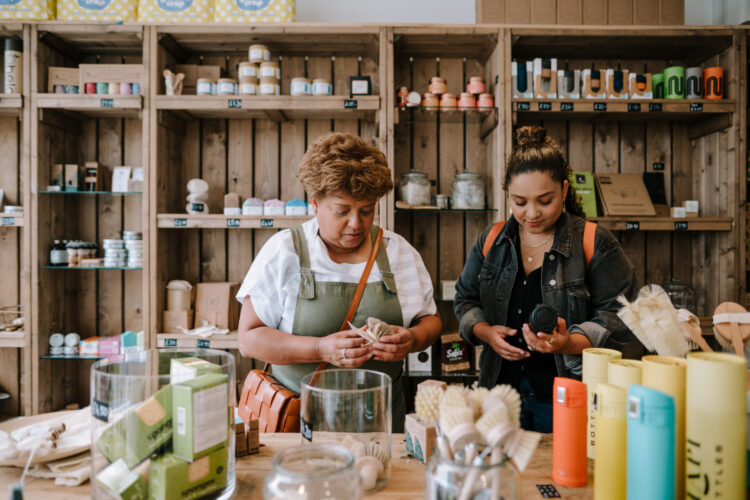 Two people are shopping. A woman is holding a brush, and a woman is holding a water bottle. There are various products stacked on wooden shelves and tables.