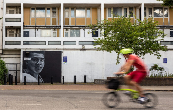 A person is cycling along a road. There is a building and a tree in the background.