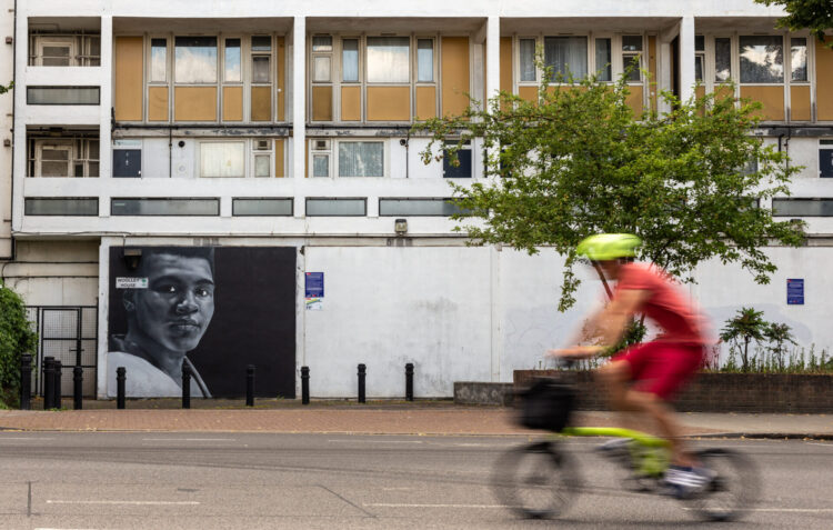 A person is cycling along a road. There is a building and a tree in the background.