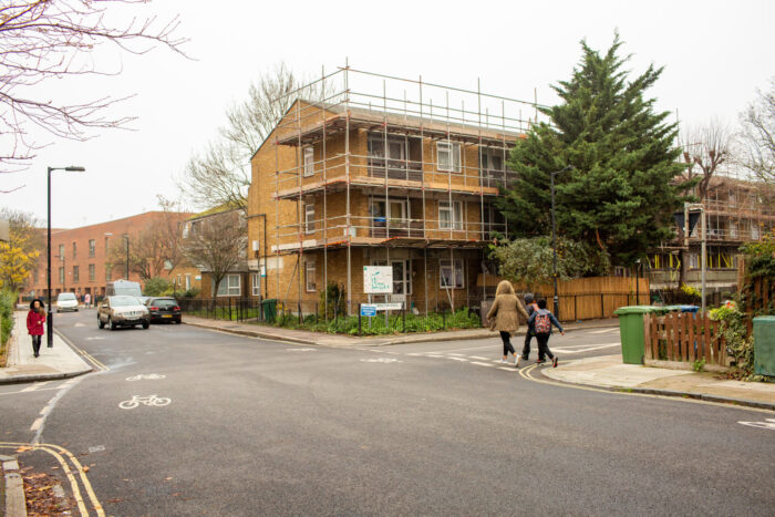 A road with a car driving down it. On the right, a building of flats and a tree are visible. Two people are walking on the pavement next to the road.