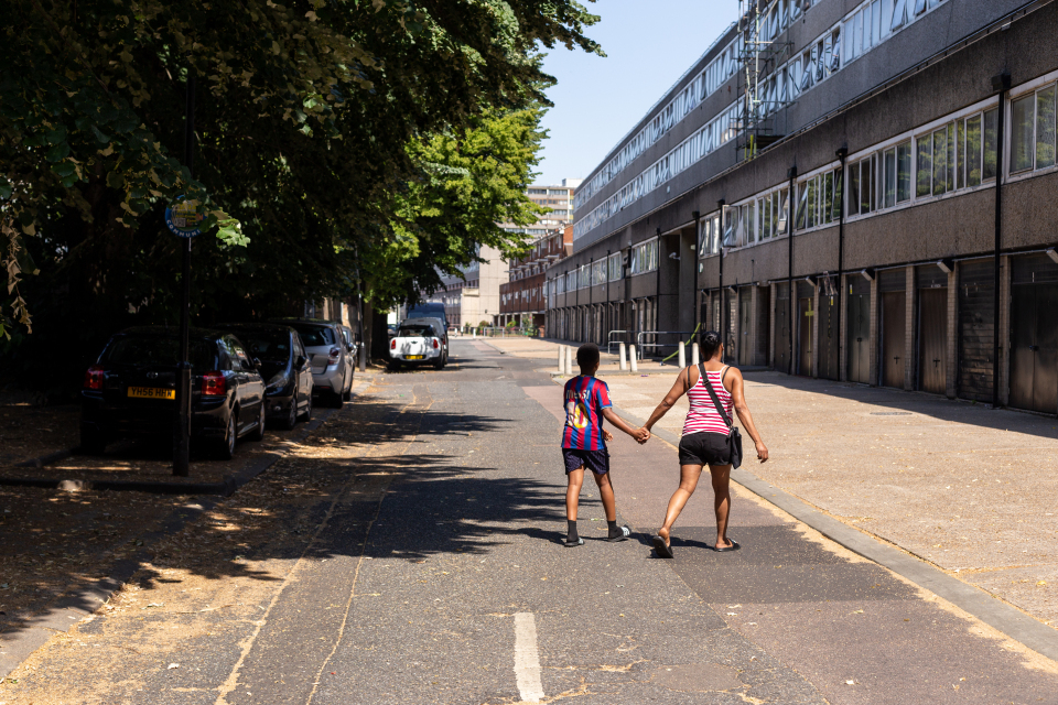 Two people are holding hands and walking down a road. Trees and a building are visible either side of the road.