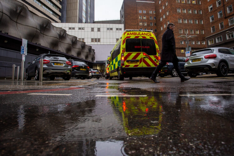 Ambulances outside a hospital. A person is walking past, and there are vehicles parked next to the ambulance.