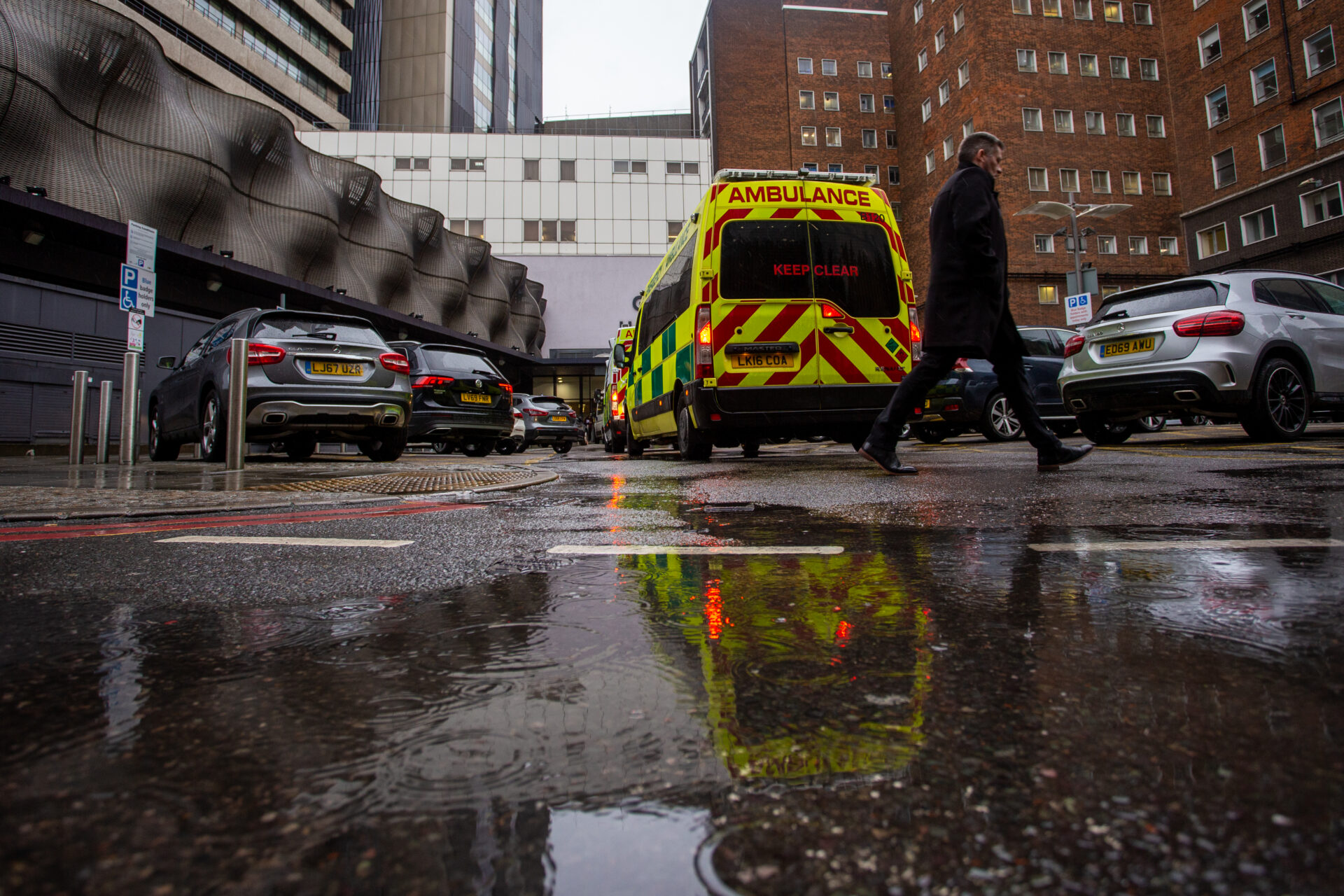 Ambulances outside a hospital. A person is walking past, and there are vehicles parked next to the ambulance.