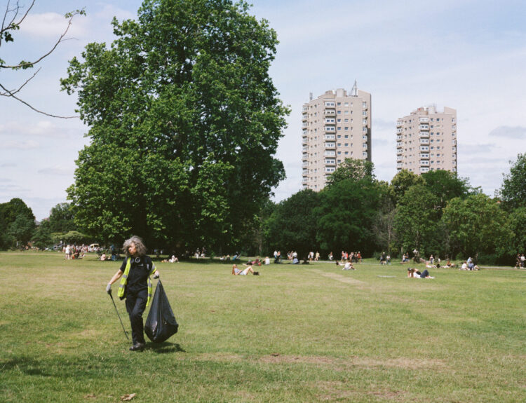 A large, grassy area with trees in the background. People are lying on the grass and walking through the park. In the foreground, a person wearing a high-vis jacket is holding a bin bag and picking up litter with a litter picker. In the distance, there are two high rise buildings.