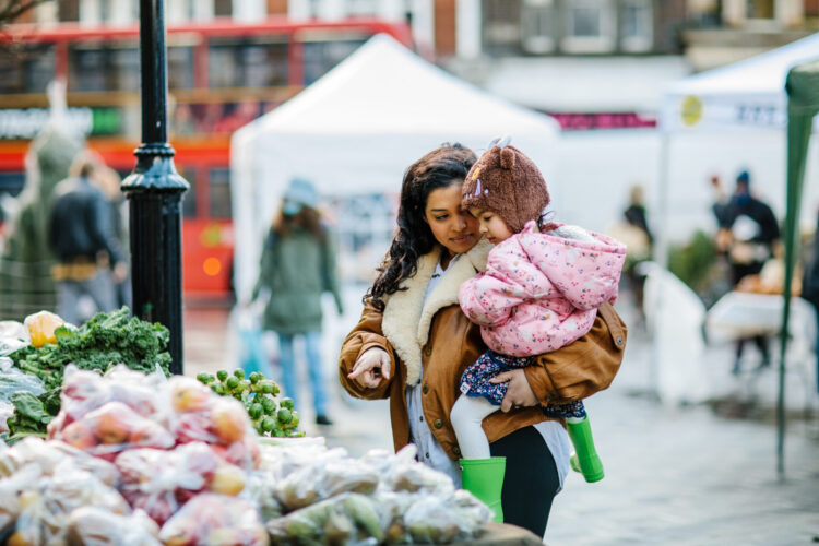 A woman is carrying a child in her arms and pointing at some vegetables on display at a market stall.