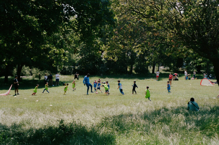 Children are playing football in a large, open and grassy space. There are trees in the background.