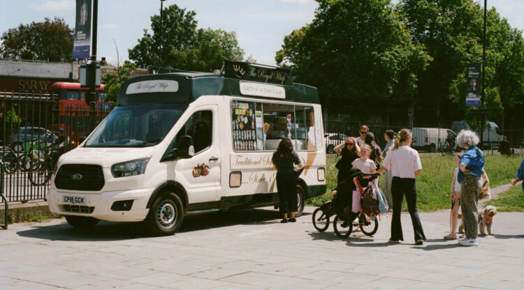 People are queuing in front of an ice cream van. There is a person with a pushchair. There is a child standing next to a dog.