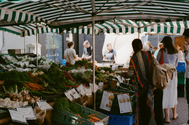 People are looking at vegetables on sale at a street market. Various vegetables, including carrots, chard, and mushrooms are on display in crates and boxes.