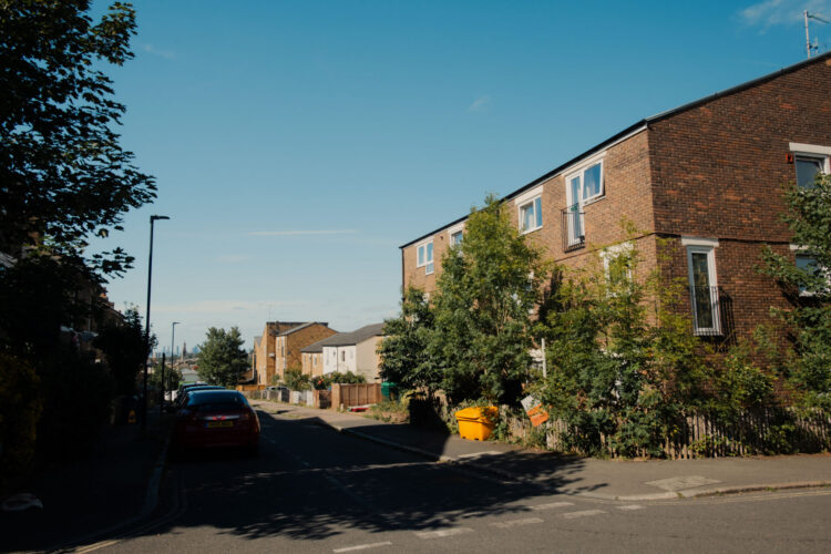 A residential street with some houses and flats. Cars are parked on the side of the road. There are trees in front of the building.
