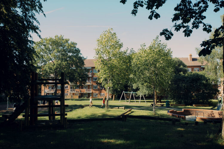 A children's playground with swings and slides. Children are running on the grass. In the distance, there are some flats.