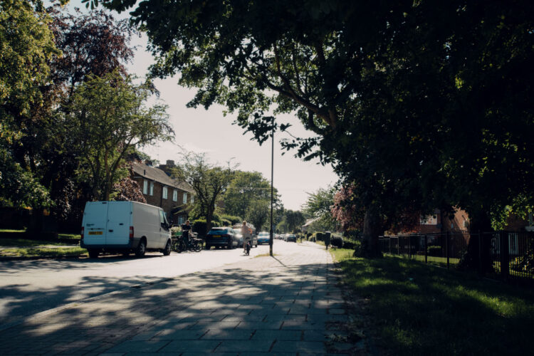 A street with houses. Vehicles and bikes are parked on the side of the road. A person is cycling down the road. There are trees next to the pavement.