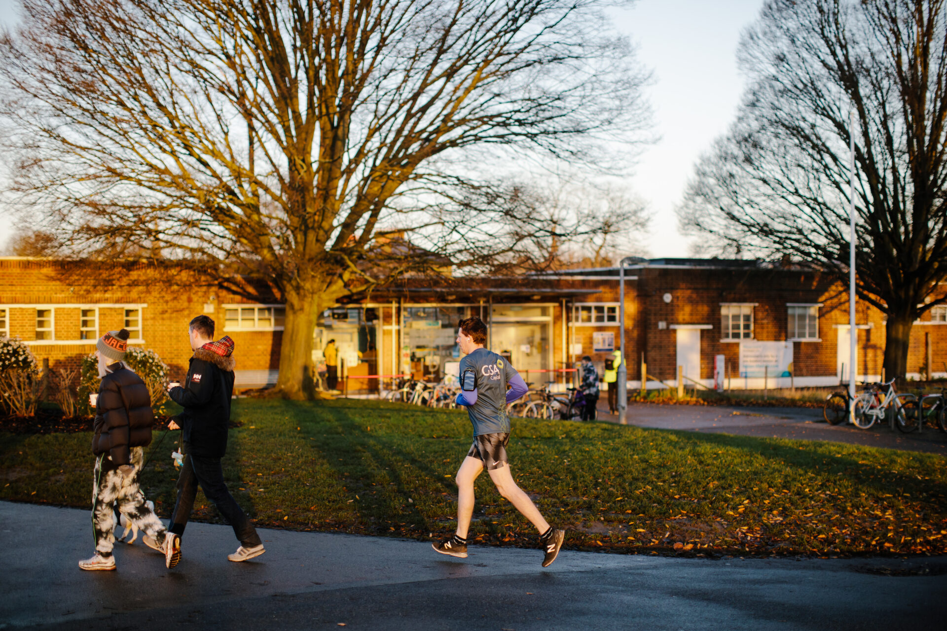 A person is jogging along a footpath. There are two people walking in front of him. In the distance, there are some buildings and a bus stop with people waiting.