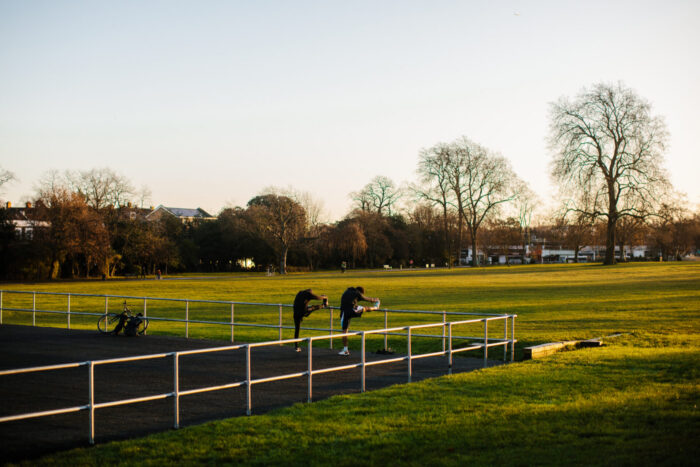 A%20field%20of%20grass%20with%20trees%20in%20the%20distance.%20Two%20people%20are%20stretching%20their%20legs%20and%20resting%20their%20feet%20on%20a%20rail.