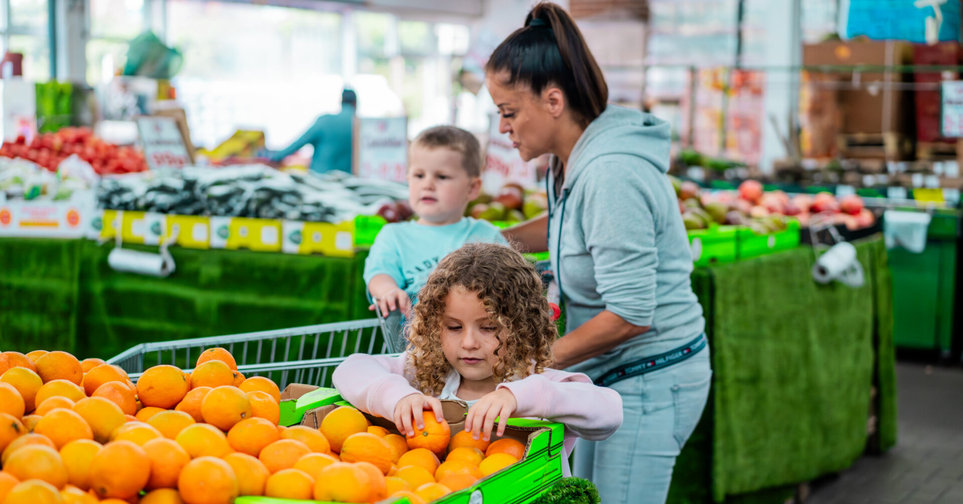 A woman is shopping for fruit with two children. One child is sitting in a seat in a shopping trolley. The other child is looking at some oranges on display.
